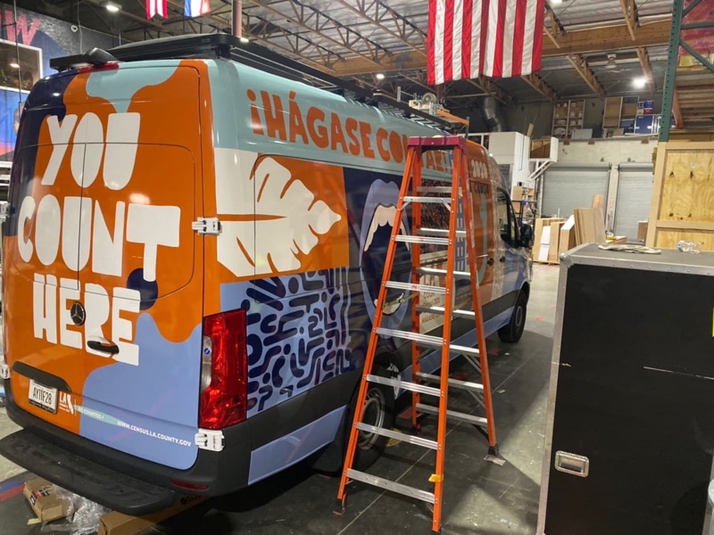 Orange and blue census outreach wrap on the rear of a sprinter van inside a wrap shop